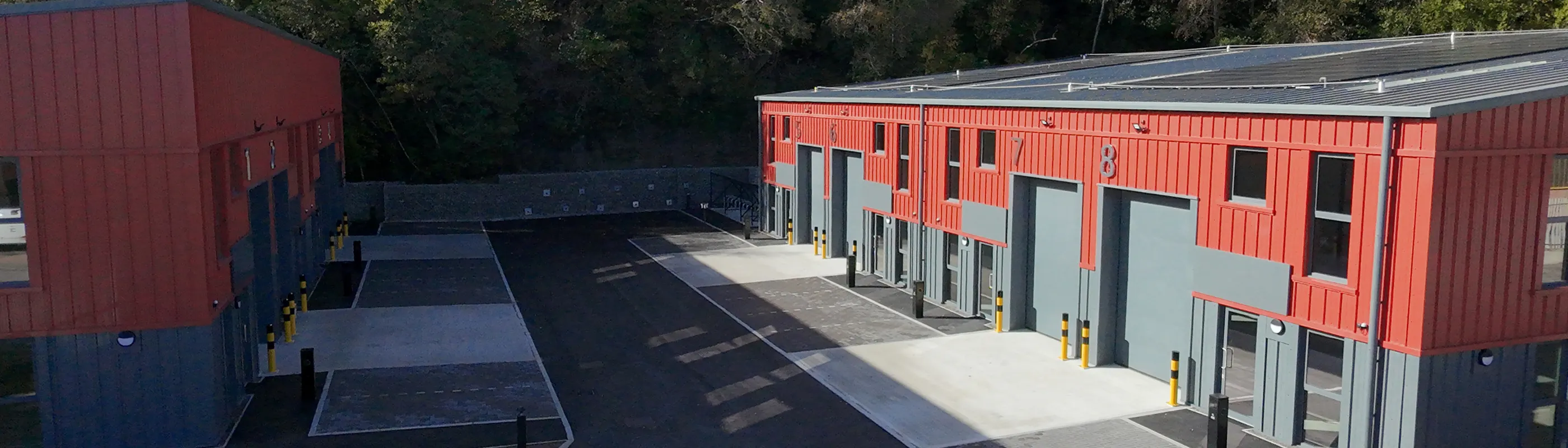 A photo of metal industrial buildings with car parking in front and trees behind. 