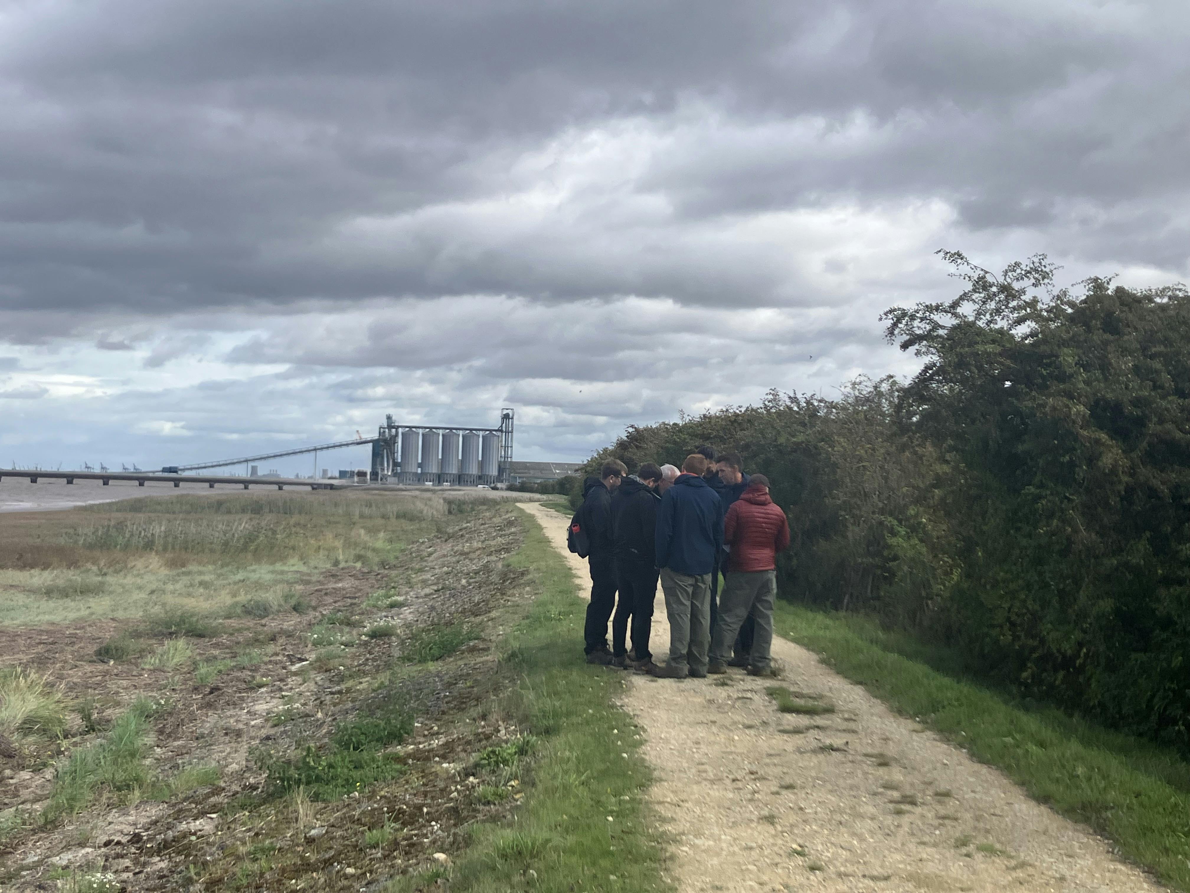 Photo of a group of people standing on a dirt path surrounded by grass and bushes, with large industrial structures and a conveyor system visible in the background under an overcast sky.