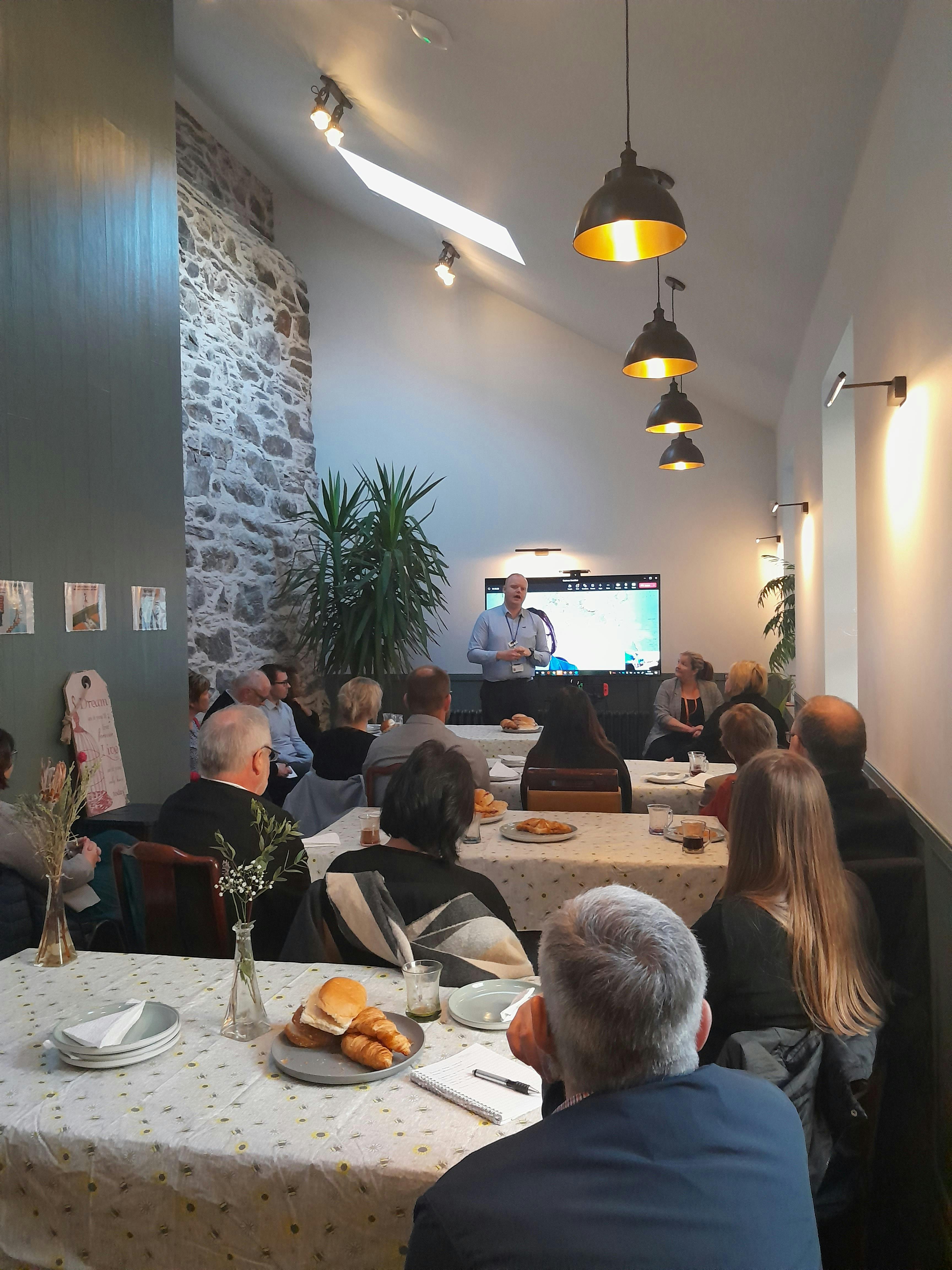 Picture shows attendees sitting at a table listening to a presentation