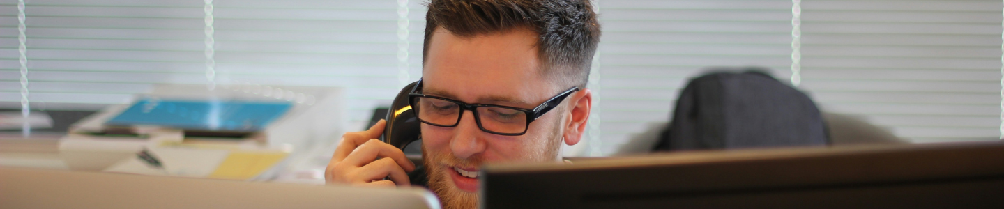 Man with glasses and a beard on phone behind computer screen