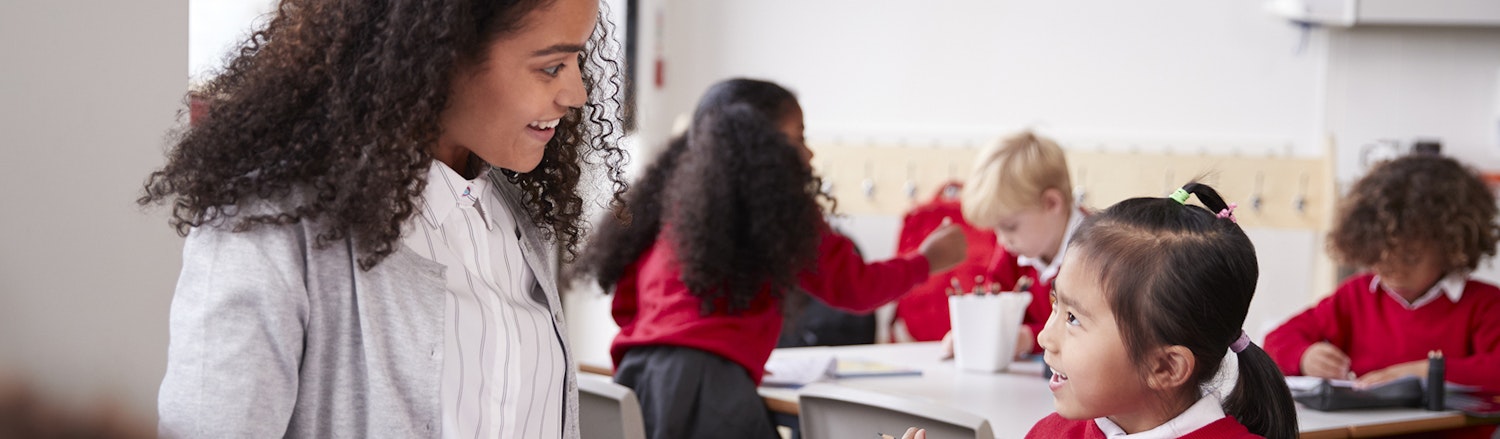 Primary school teacher sitting at table in a classroom talking to a young schoolgirl
