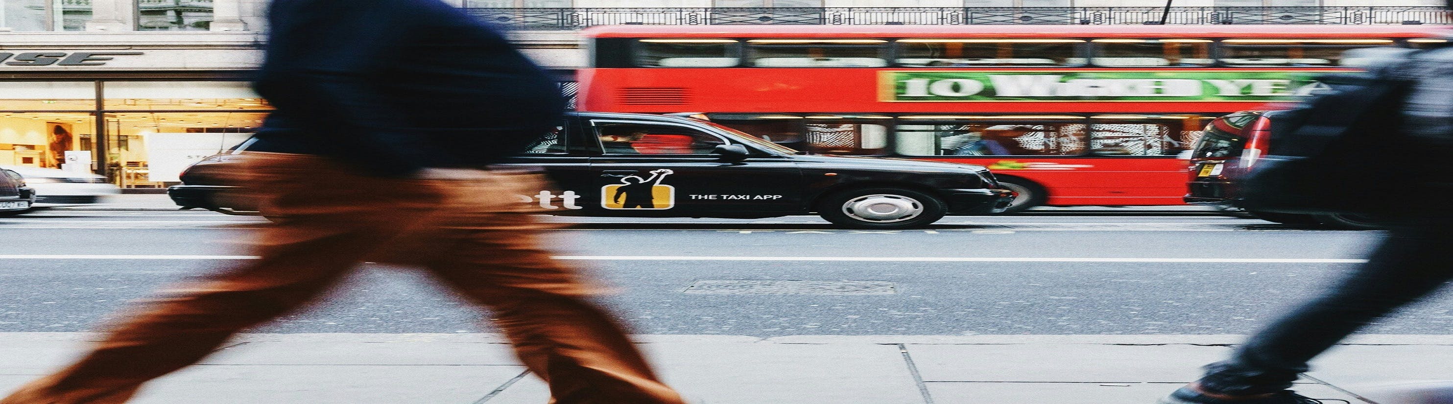 Photo of people walking with black cab & red bus in background.