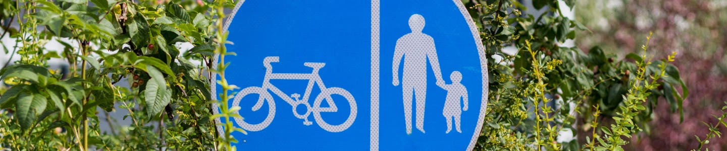 Image of a blue circular sign with an icon of a bike on the left and an adult and child on the right, surrounded by foliage.