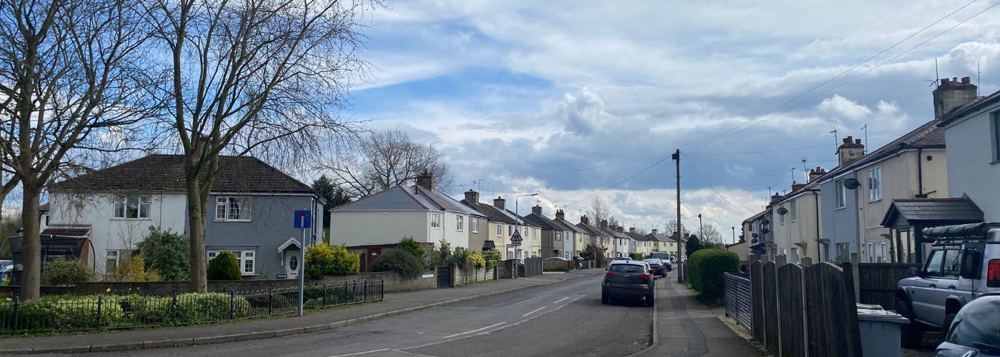 A street of houses with trees at the end and cars parked on the side of the road.