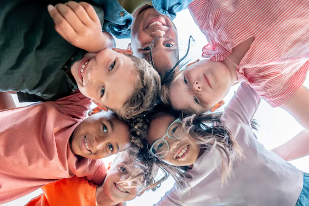 Six children in a circle, with arms around each other, looking downwards towards the camera